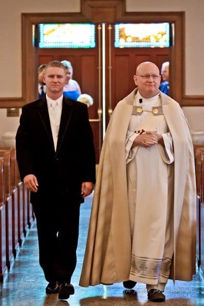raleigh wedding photographer - jenna and ryan A man in a black suit walks alongside a clergyman in white and beige robes down the aisle of a church, with wooden pews on either side. Stained glass windows are visible in the background.