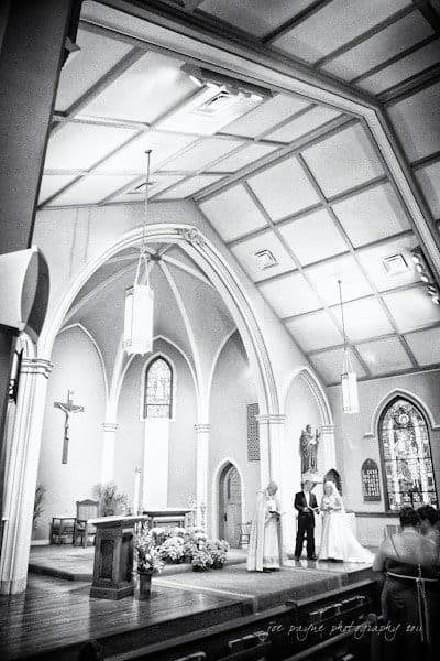 raleigh wedding photographer - jenna and ryan Black and white photo of a church interior featuring an arched ceiling and stained glass windows. A couple stands at the altar with an officiant. The church is decorated simply, with a large cross on the wall.