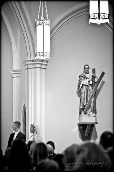 raleigh wedding photographer - jenna and ryan Black and white image of a wedding ceremony inside a church. A bride and groom stand together beneath Gothic architecture, near a statue of a saint holding a cross. Tall lanterns hang from the ceiling, adding to the solemn atmosphere.