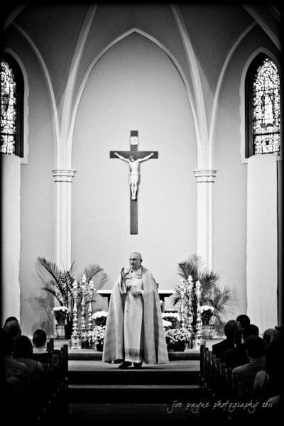 raleigh wedding photographer - jenna and ryan A black-and-white photo of a church interior, featuring a priest standing in front of an altar. A large crucifix hangs on the wall behind him. Parishioners are seated in pews, and the space is adorned with plants and flowers.