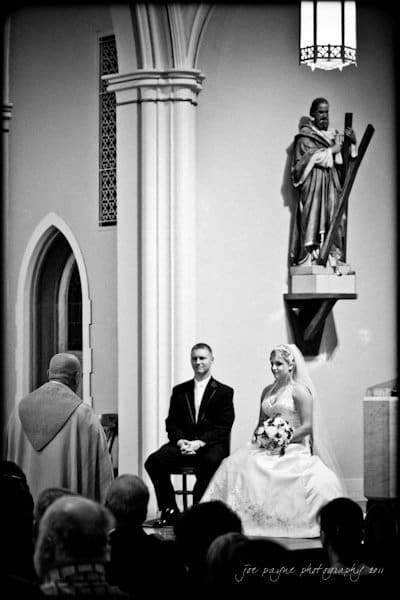 raleigh wedding photographer - jenna and ryan A bride and groom sit facing a religious officiant in a church. The bride wears a white gown and veil, holding a bouquet, while the groom is in a dark suit. A statue is visible in the background. The atmosphere is solemn and formal.