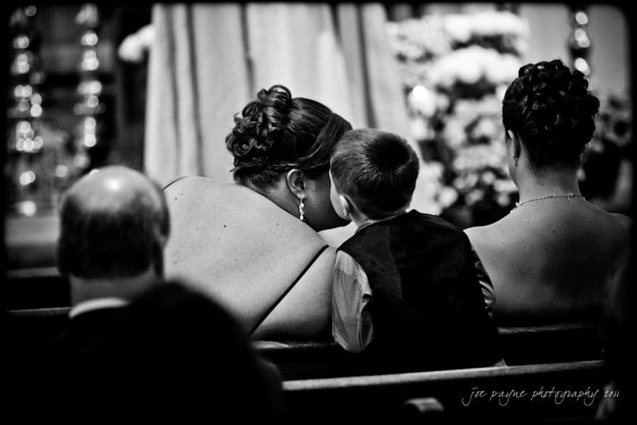 raleigh wedding photographer - jenna and ryan A black and white photo of a woman and a young boy sitting on a pew at a wedding ceremony. The boy is whispering to the woman, who is looking toward the front. Another woman is seated next to them. Decorations are visible in the background.