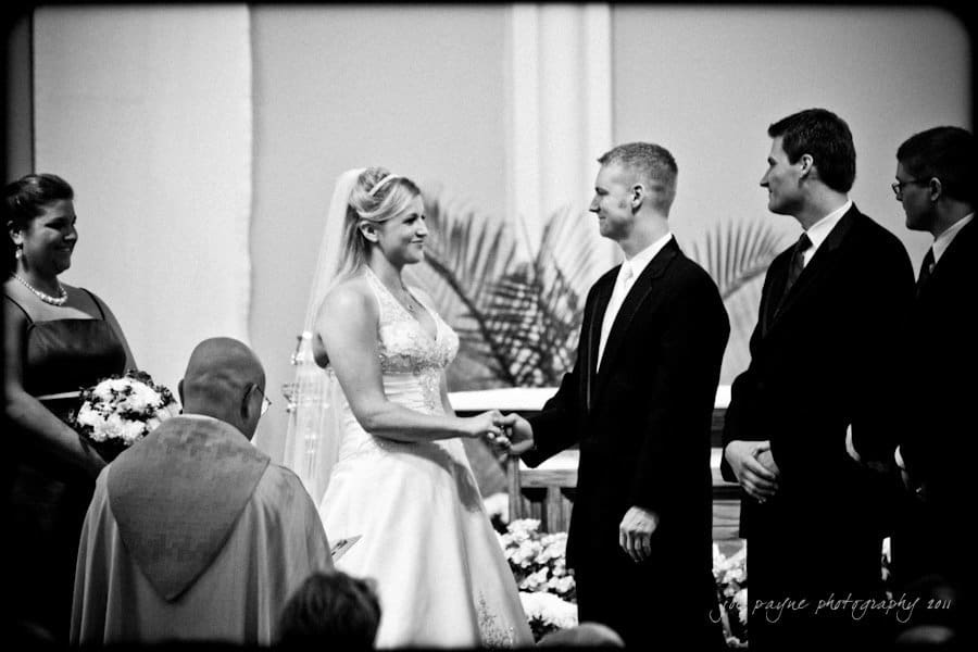 raleigh wedding photographer - jenna and ryan A black and white photo of a bride and groom holding hands and smiling at each other during a wedding ceremony. The officiant and two groomsmen stand nearby, and a bridesmaid holds a bouquet.