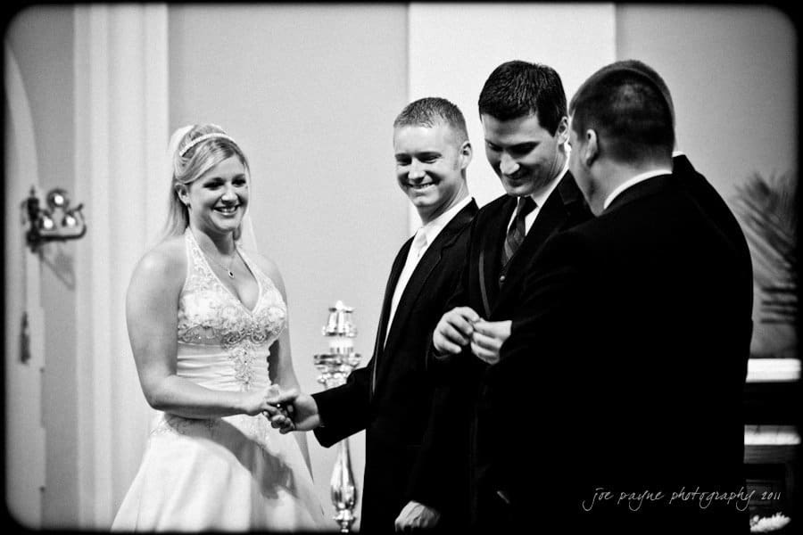 raleigh wedding photographer - jenna and ryan A black and white photo of a bride and groom holding hands, smiling, as two groomsmen stand beside them. They are inside a building with arches, possibly a church, during a wedding ceremony.