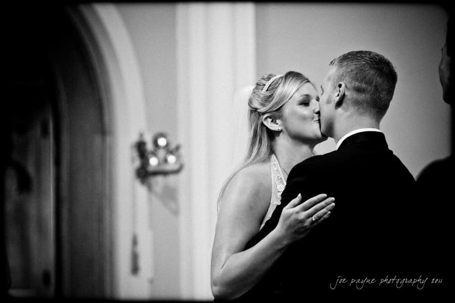 raleigh wedding photographer - jenna and ryan A black and white image of a couple kissing during their wedding ceremony. The bride wears a halter dress with an updo hairstyle, while the groom is in a suit. Soft lighting and a blurred background create an intimate atmosphere.