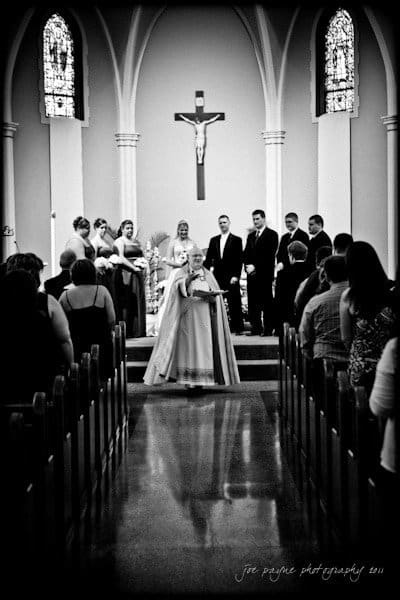 raleigh wedding photographer - jenna and ryan A black and white photo of a wedding ceremony in a church. A priest stands at the altar with a bridal couple and attendants. Guests are seated and watching the ceremony. A crucifix and stained glass windows are visible in the background.