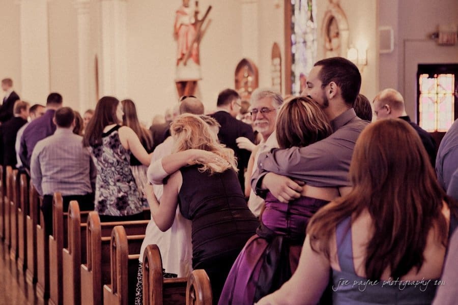raleigh wedding photographer - jenna and ryan People in a church exchanging hugs and handshakes during a service. The scene is warm and conveys a sense of community. Stained glass windows and wooden pews are visible in the background.