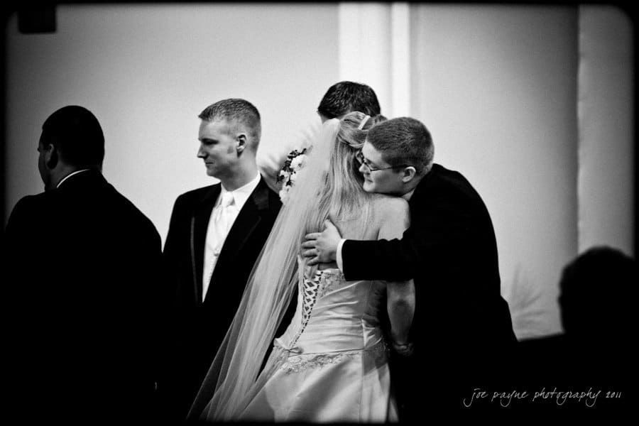 raleigh wedding photographer - jenna and ryan A black and white image of a bride in a wedding gown hugging a man wearing a suit. Theyre surrounded by other suited men, seemingly at a wedding ceremony.