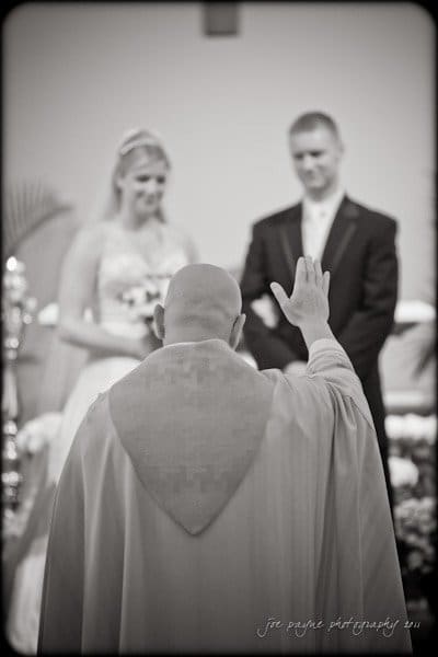 raleigh wedding photographer - jenna and ryan A black and white photo of a wedding ceremony. A couple stands together, holding hands. A priest with a raised hand is blessing them. The bride is in a gown and the groom in a suit.