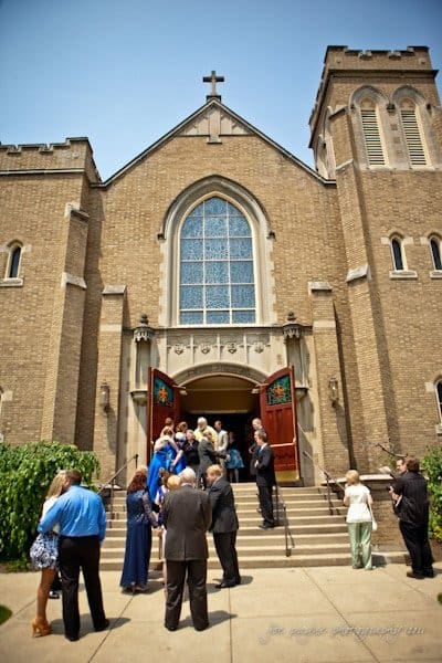 raleigh wedding photographer - jenna and ryan A group of people stand and converse on the steps leading to the open double doors of a large brick church. The building has tall windows and a cross on top. The sky is clear and bright.