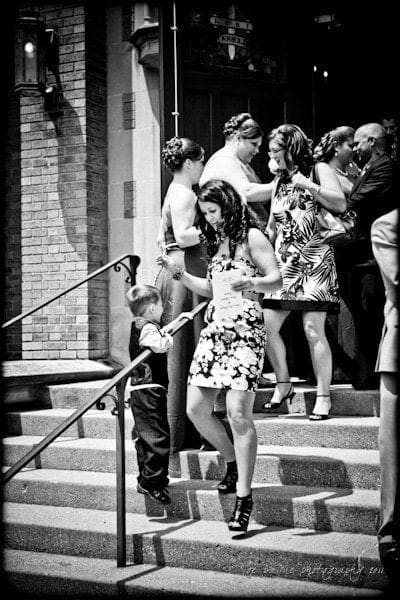 raleigh wedding photographer - jenna and ryan A black and white photo shows a group of adults and a child on steps outside a brick building. The child, in formal attire, holds hands with a woman in a floral dress. Others nearby are in conversation, dressed in various patterned outfits.
