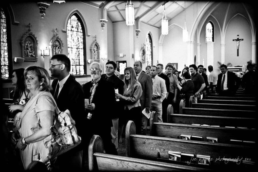 raleigh wedding photographer - jenna and ryan A black and white photo of people standing in line inside a church. The church features stained glass windows and wooden pews. The group appears to be attending an event or service.