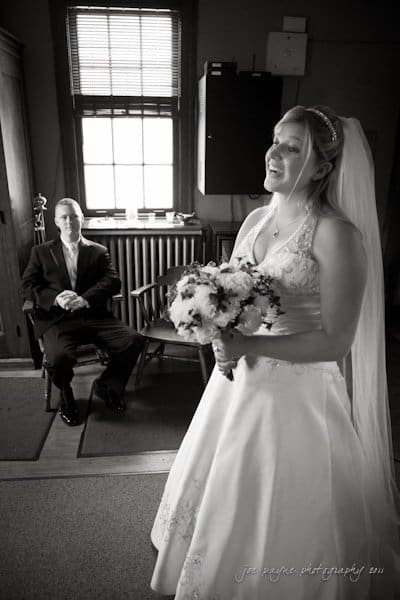 raleigh wedding photographer - jenna and ryan A bride in a wedding gown holds a bouquet, smiling, as she stands near a window. A man in a suit sits in the background, watching her with a calm expression. The room is softly lit, creating a serene atmosphere.
