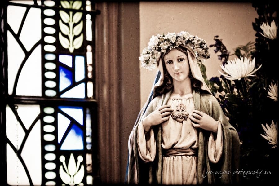 raleigh wedding photographer - jenna and ryan A statue of the Virgin Mary adorned with a floral crown is positioned beside an intricately patterned stained glass window. The statues hands gently touch her chest, and the setting includes white flowers in the background.