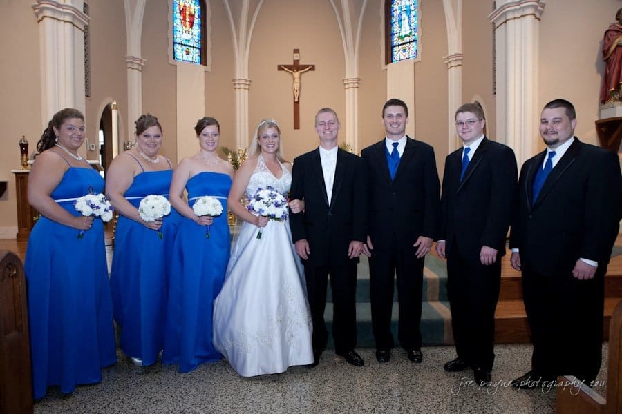 raleigh wedding photographer - jenna and ryan A bridal party in a church with stained glass windows. The bride is in a white gown, holding blue and white flowers. She is surrounded by bridesmaids in blue dresses and groomsmen in black suits with blue vests. A crucifix is visible in the background.