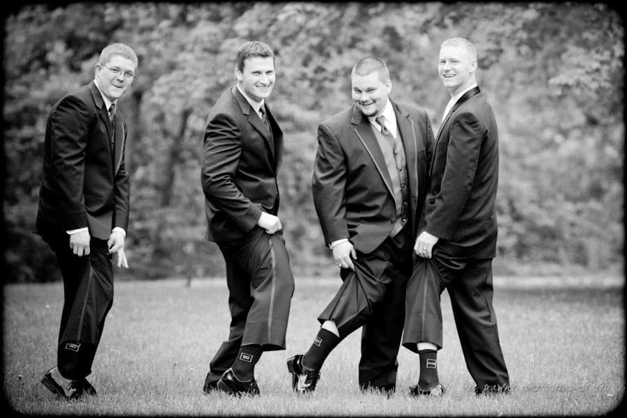 raleigh wedding photographer - jenna and ryan Four men in suits, standing outdoors, smiling and showing off their socks. The image is in black and white, with a background of trees and grass.