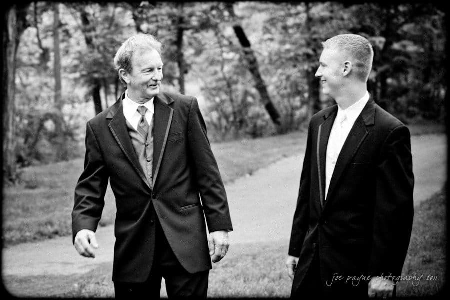 raleigh wedding photographer - jenna and ryan Black and white photo of two men in suits walking and smiling at each other. They appear to be in a park setting with trees and a path in the background.