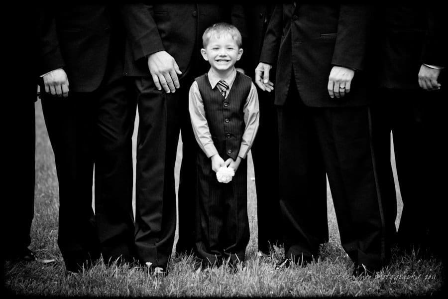 raleigh wedding photographer - jenna and ryan A young boy in a suit stands smiling among a group of adults in formal attire. The adults faces are not visible, and the focus is on the boy holding a small object, standing on grass. The image is in black and white.