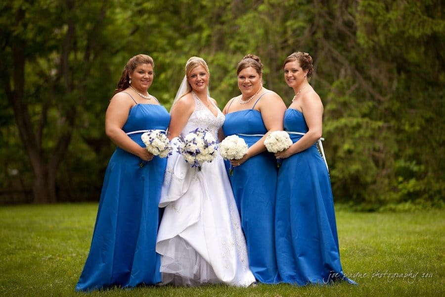 raleigh wedding photographer - jenna and ryan Four women in a grassy area, with the woman in the center wearing a white bridal gown and holding a bouquet of white and blue flowers. The three other women are in matching blue dresses, each holding a white bouquet. Green trees are in the background.