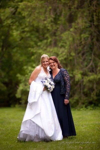 raleigh wedding photographer - jenna and ryan A bride in a white gown holding a bouquet is smiling and standing next to a woman in a dark blue dress. They are outdoors on a grassy area with lush green trees in the background.