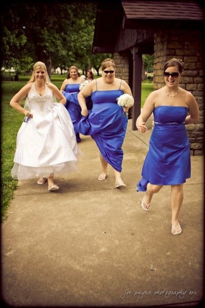 raleigh wedding photographer - jenna and ryan A bride in a white dress and three bridesmaids in blue dresses are joyfully running on a park path. The bridesmaids are holding their dresses and one has a bouquet. Trees and a stone building feature in the background.