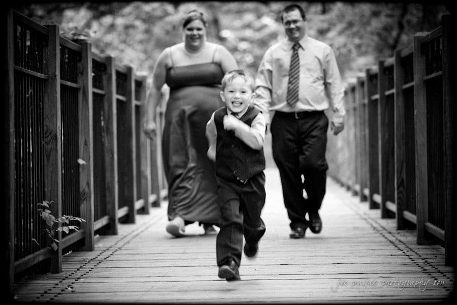 raleigh wedding photographer - jenna and ryan A young boy in a vest and dress shirt runs joyfully on a wooden bridge. Behind him, a man in a shirt and tie and a woman in a long dress walk together, smiling. The scene is in black and white.