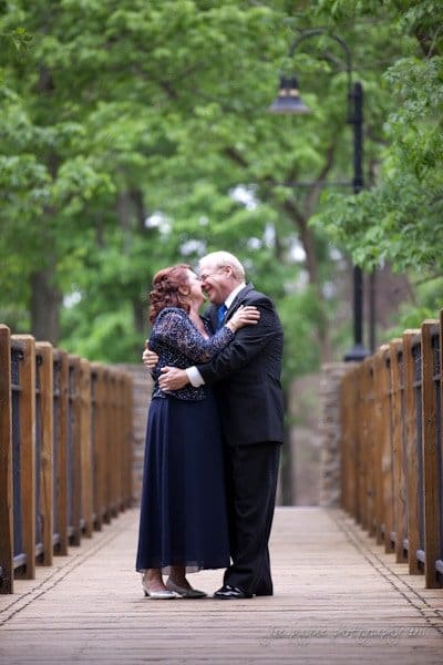 raleigh wedding photographer - jenna and ryan An elderly couple stands embracing on a wooden bridge surrounded by greenery. They are dressed formally and appear joyful, sharing a tender moment. The bridge is lined with rustic railings and leads into a lush forest path.