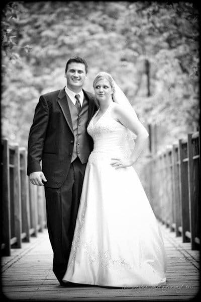 raleigh wedding photographer - jenna and ryan A black and white photo of a bride and groom standing on a wooden bridge. The groom is in a suit, and the bride is wearing a wedding dress with a veil. Both are smiling and posing together, surrounded by trees in the background.