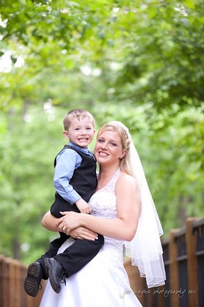 raleigh wedding photographer - jenna and ryan A smiling bride in a white dress and veil holds a young boy wearing a blue shirt and black vest. They stand outdoors on a wooden pathway surrounded by lush green trees.
