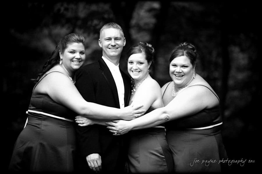 raleigh wedding photographer - jenna and ryan A black and white photo of a smiling man in a suit surrounded by three women in matching dresses. The women have their arms around him, and they all appear happy, posing outdoors.
