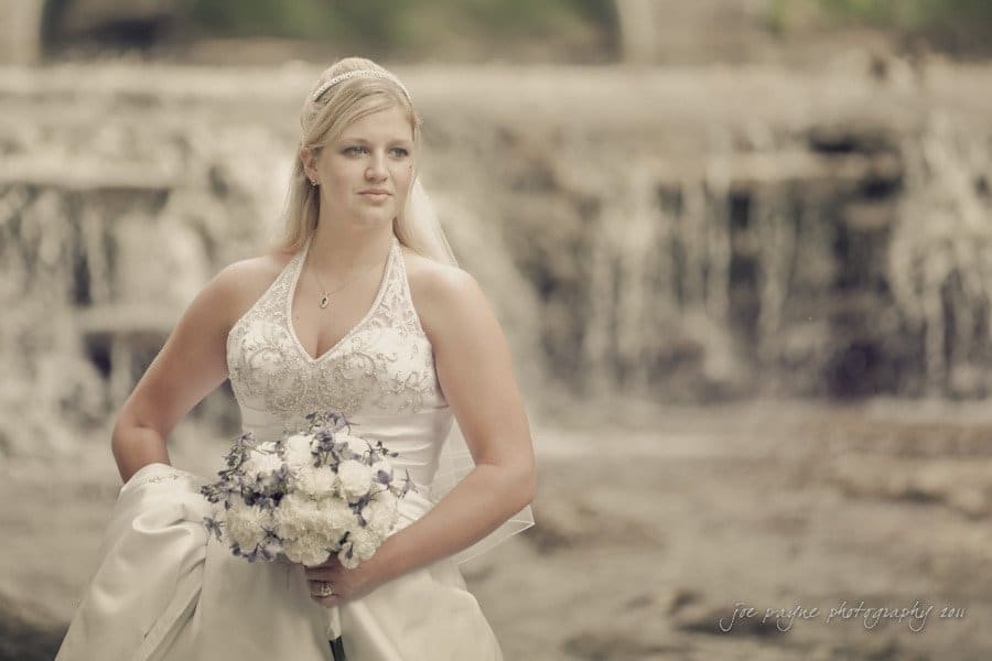 raleigh wedding photographer - jenna and ryan A bride stands outdoors in front of a waterfall, wearing a white gown with intricate details. She holds a bouquet of white and purple flowers and gazes into the distance. Her veil is attached to her styled blonde hair.