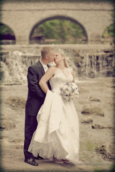 raleigh wedding photographer - jenna and ryan A couple in wedding attire shares a tender moment by a rocky riverbank. The bride holds a bouquet of white and blue flowers, while the groom embraces her from behind. In the background, an arched stone bridge spans across the river.