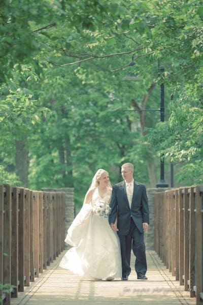 raleigh wedding photographer - jenna and ryan A bride in a white gown and a groom in a black suit walk hand in hand on a wooden bridge surrounded by lush green trees. The sunlight filters through the leaves, creating a serene and romantic atmosphere.