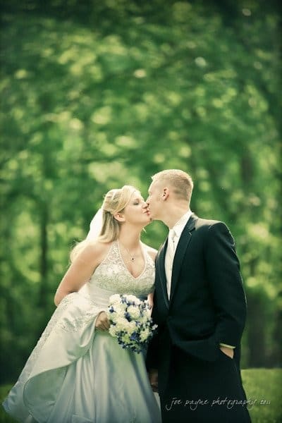 raleigh wedding photographer - jenna and ryan A bride in a white gown and a groom in a dark suit share a kiss outdoors. She holds a bouquet with white and purple flowers. The background is lush and green, suggesting a forest or park setting.