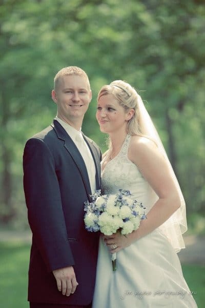 raleigh wedding photographer - jenna and ryan A bride and groom stand together outdoors. The groom is dressed in a dark suit, and the bride wears a white gown and veil, holding a bouquet of white and purple flowers. The background is a blurred, lush green setting.