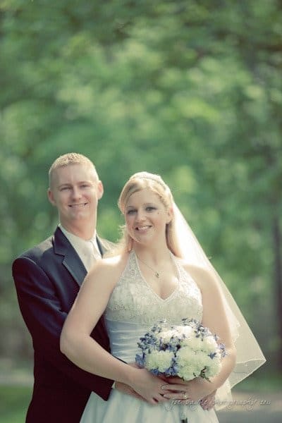 raleigh wedding photographer - jenna and ryan A bride and groom stand closely together outdoors, with the groom in a black suit and the bride in a white gown holding a bouquet of white and purple flowers. Lush green trees are in the background, creating a soft, serene setting.