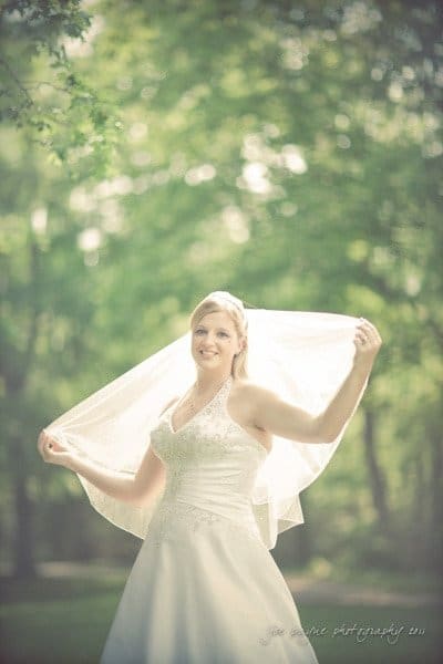 raleigh wedding photographer - jenna and ryan A bride in a white wedding dress holds her veil above her shoulders, standing outdoors with a soft focus background of green trees. Sunlight filters through the leaves, creating a dreamy, serene atmosphere.
