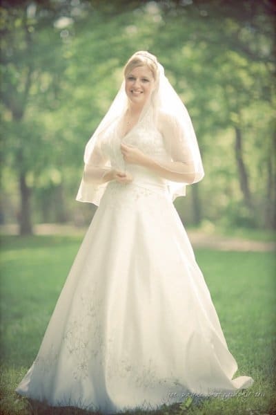 raleigh wedding photographer - jenna and ryan A bride stands smiling in a sunlit outdoor setting, wearing a white wedding dress and veil. She poses gracefully on green grass with trees blurred in the background, creating a serene and joyful atmosphere.