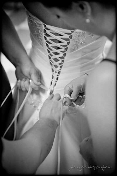 raleigh wedding photographer - jenna and ryan Three people are tying the corset back of a wedding dress, focusing on the intricate lacing. The brides gown features delicate embroidery. The image is in black and white, emphasizing the details of the dress and laces.