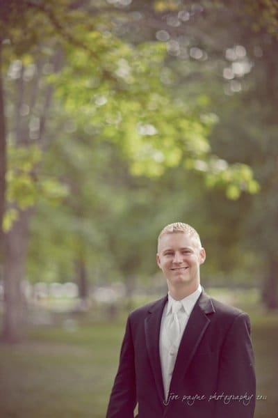 raleigh wedding photographer - jenna and ryan A person in a suit smiles while standing outdoors in a park with blurred green trees in the background.