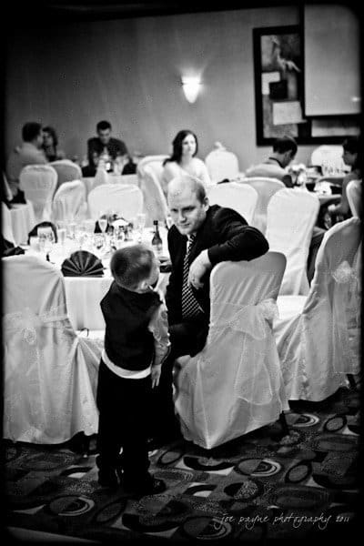 raleigh wedding photographer - jenna and ryan A young boy in a suit talks to a seated man at a decorated banquet table in a dimly lit reception room. Other guests sit at tables in the background.