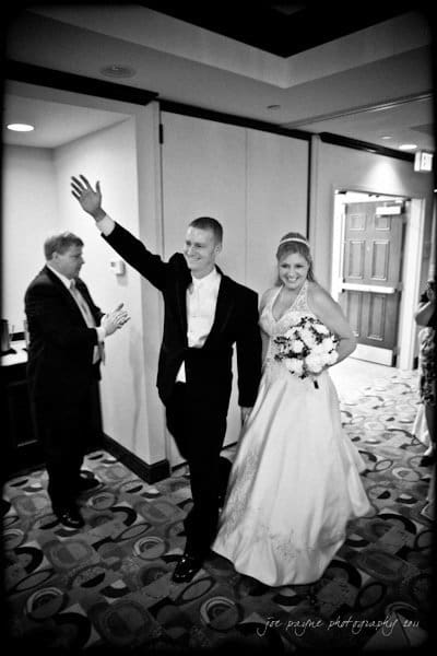 raleigh wedding photographer - jenna and ryan A bride and groom walk arm in arm down a hallway. The groom waves to the side while the bride holds a bouquet of flowers. They are smiling and dressed in formal wedding attire. A man stands nearby clapping for them.