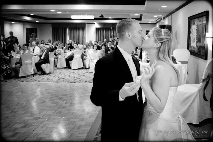 raleigh wedding photographer - jenna and ryan A bride and groom share a kiss on the dance floor at their wedding reception. Guests sit at decorated tables in the background, watching the couple. The setting is a warmly lit banquet hall.