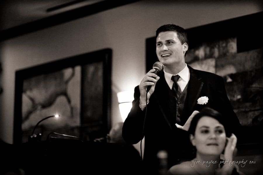 raleigh wedding photographer - jenna and ryan A man in a suit, holding a microphone, smiles as he speaks in a dimly lit room. A woman in the foreground sits and listens. The background features abstract artwork on the walls.