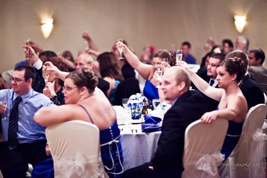 raleigh wedding photographer - jenna and ryan A group of people in formal attire raising glasses for a toast at a wedding reception. The room is decorated elegantly, and guests are seated around tables with white tablecloths and blue floral centerpieces.