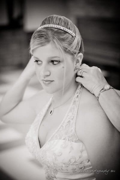raleigh wedding photographer - jenna and ryan A bride in a white gown adjusts her necklace with the help of someone. She has an intricate headband in her hair and appears to be in a softly lit room. The image is in black and white.