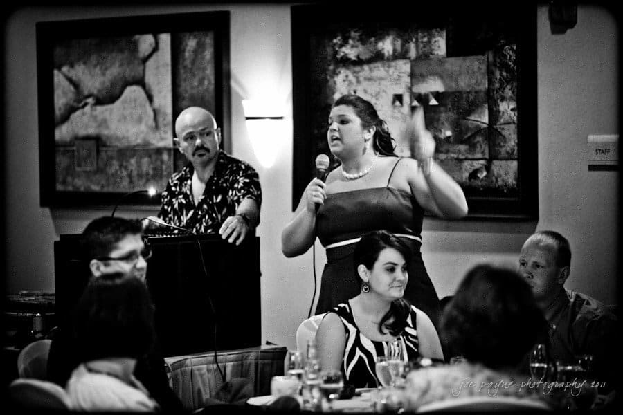 raleigh wedding photographer - jenna and ryan A black and white photo of a woman speaking into a microphone at an event, with a man standing at a podium behind her. Guests are seated at tables listening. The room is decorated with abstract paintings.