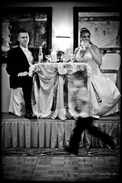 raleigh wedding photographer - jenna and ryan A bride and groom sit at a decorated table, looking towards the left. A child in motion blurs past in the foreground. The black and white setting creates a classic, formal atmosphere.