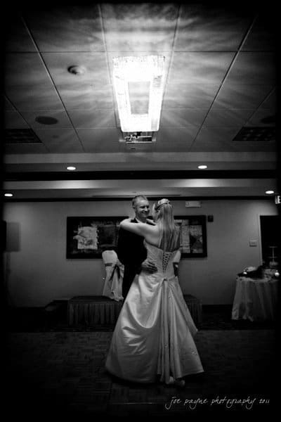 raleigh wedding photographer - jenna and ryan A black and white photo of a couple dancing in an elegant room. The woman is wearing a long gown, and the man is in a suit. They are smiling at each other under a central ceiling light, creating a warm and intimate atmosphere.