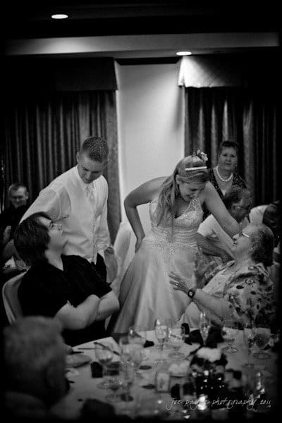 raleigh wedding photographer - jenna and ryan A bride in a wedding dress interacts playfully with a seated guest, while a groom in a vest stands beside her, smiling. Other guests are seated at a table with glasses and plates. The mood is joyful and lively in a warmly lit room.